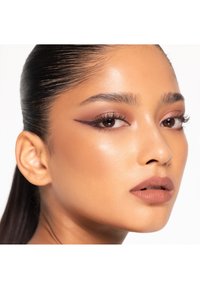 Nahaufnahme eines Gesichts mit glatter Haut, definierten Augenbrauen, kräftigem Eyeliner und matten mauvefarbenen Lippen. Die Haare sind zurückgekämmt und sehen nass aus.