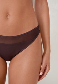 Brązowe bikini z przezroczystym siateczkowym paskiem w talii, o niskim kroju i gładkiej fakturze. Bez widocznych wzorów ani detali metalowych.