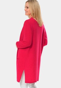 Gilet long en tricot rouge avec une coupe décontractée, fentes latérales et poches avant. Texture douce, avec des manches longues et un design ouvert à l'avant.