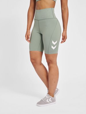 Kvinde iført salviegrøn cykelshorts med høj talje med hvide chevron-logoer og matchende grå sneakers med hvide snørebånd og chevron-logoer.