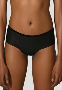 Kvinna som bär enkla svarta sömlösa hipster-undertøj, visad från midjan till övre låren mot en ljus bakgrund.