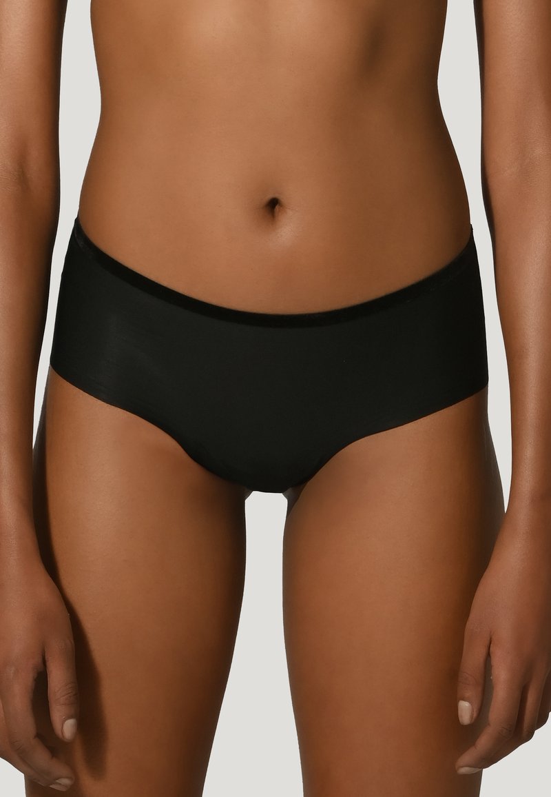 Kvinna som bär enkla svarta sömlösa hipster-undertøj, visad från midjan till övre låren mot en ljus bakgrund.