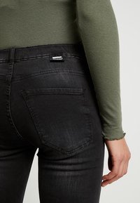 Jeans pitillo negros con un acabado texturizado y un sutil desgaste, con un bolsillo trasero y una pequeña etiqueta de la marca. Combinados con un top verde de punto acanalado.