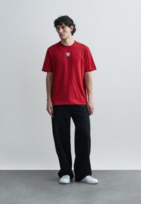 Camiseta de algodón roja con cuello redondo, mangas cortas y un estampado gráfico en la parte delantera. Combinada con pantalones negros de corte ancho y zapatillas blancas.