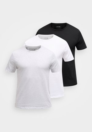 Drie eenvoudige Lacoste crewneck T-shirts in lichtgrijs, wit en zwart, gerangschikt in lagen van voren naar achteren op een witte achtergrond.