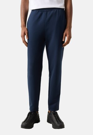 Mann trägt marineblaue Slim-Fit-Hose mit elastischem Bund und schwarze Freizeitsneaker, steht vor einem hellen Hintergrund.