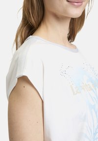 White short-sleeve shirt with blue floral design and gold text. Features grey accents on neckline and shoulders, with rhinestone embellishments.