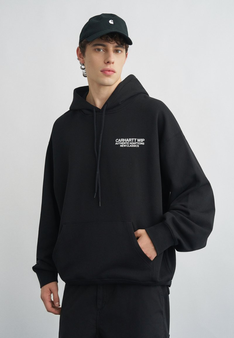 Giovane uomo che indossa una felpa nera Carhartt WIP con cappuccio e un cappellino nero, in piedi con una mano nella tasca della felpa contro uno sfondo semplice.