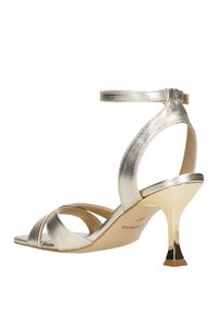 Sandaletta a strappo alla caviglia di colore oro metallico con design incrociato, punta affusolata e tacco a stiletto con base quadrata. Texture liscia.