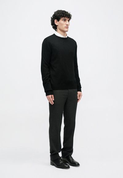 Un pull noir à col rond avec des poignets côtelés, associé à un pantalon tailleur gris foncé et des chaussures en cuir noires, sur un fond blanc uni.