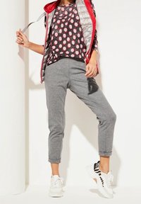 Rotes und graues gemustertes Oberteil, graue, cropped Hosen und weiße Sneakers mit schwarzen Streifen. Die Weste ist silberfarben und hat eine glatte Textur.