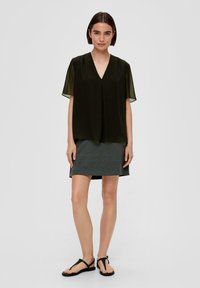 Schwarzes transparentes Oberteil mit kurzen Ärmeln, V-Ausschnitt, über einem grauen Rock. Modell trägt schwarze flache Sandalen und steht vor einem neutralen Hintergrund.