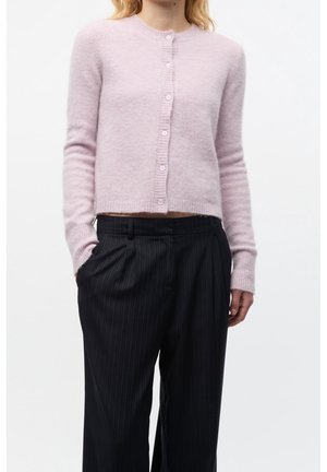 Žena v světle růžovém chlupatém kardiganu na knoflíky a černých širokých kalhotách s proužky, ruce v kapse, stojící před bílým pozadím.