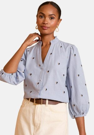 Donna che indossa una blusa a strisce blu chiaro con piccoli motivi a cuore, pantaloni crema, cintura marrone e orecchini a cerchio, mentre si tocca il collo.
