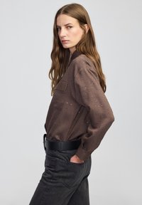 Bruin gestreept shirt met lange mouwen en subtiele versieringen, gecombineerd met donkergrijze jeans. Het shirt heeft een enkele borstzak en een losvallende pasvorm.