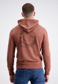 Bruine hoodie met een trekkoord en een logoprint op de achterkant. De stof lijkt zacht, met een geribde zoom en manchetten. Gemaakt in combinatie met blauwe jeans.