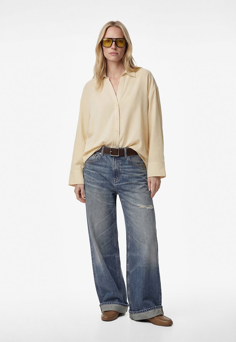 Persona rubia con gafas tintadas de amarillo, camisa beige holgada abotonada, vaqueros anchos desgastados con rotura, cinturón marrón y zapatos marrones, de pie.