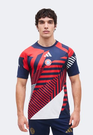 Młody mężczyzna w czerwonej, granatowej i białej koszulce piłkarskiej FC Bayern Monachium z grafiką oraz granatowych spodniach z odpowiednimi logo.