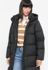Vrouw in een zwarte pufferjas met een capuchon, beige en witte gestreepte trui, witte shirt en blauwe jeans met een zwarte riem, kijkt naar links.