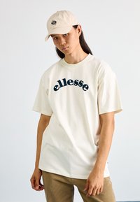 Camiseta de algodón color beige con el logo "ellesse" en azul marino oscuro, de corte holgado, con cuello redondo, combinada con una gorra beige y pantalones de pana marrón claro.