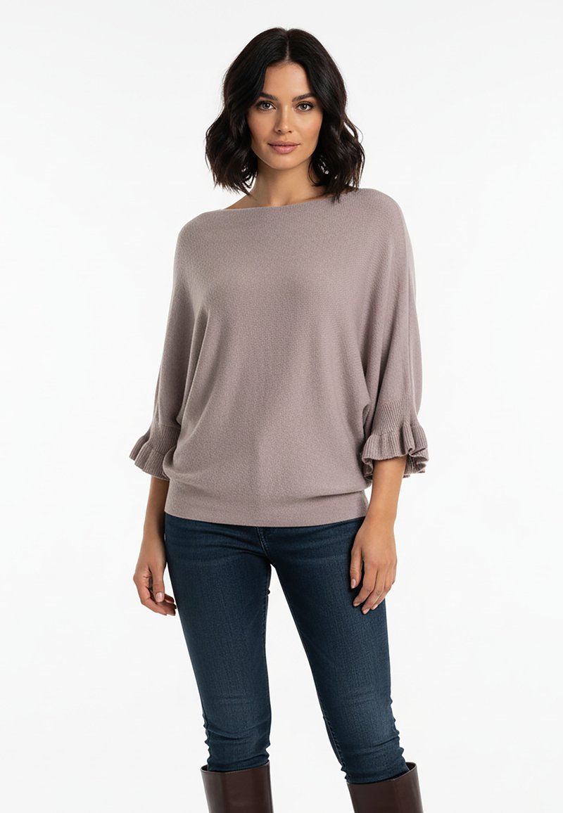 Jasnoszary, dziergany sweter o fakturze z szerokim dekoltem typu łódka i marszczonymi mankietami, zestawiony z ciemnoniebieskimi obcisłymi jeansami i brązowymi butami.