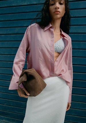 Jeune femme portant une chemise ample rose rayée par-dessus un soutien-gorge à motifs, une jupe blanche, tenant un sac à main marron texturé, devant un mur bleu.
