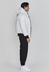Hvid pufferjakke med hætte, med sorte snoretræk og lynlås foran. Sammen med sorte sweatpants og hvide sneakers.