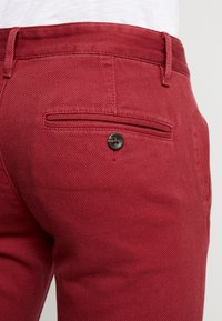 Close-up van rode textuur broek met een achterzak met zwarte knop en riemlussen, gedragen met een witte top.