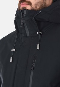 Giacca nera con colletto alto, dotata di cerniere e cordini. La texture appare liscia con accenti metallici sui dettagli.