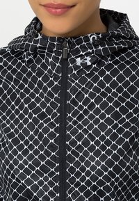 Close-up of person wearing black hooded jacket with white diamond pattern and Under Armour logo, zipper fully closed.