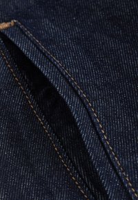 Țesătură din denim albastru închis cu un model fin de urzeală și bătătură, prezentând o cusătură vizibilă cu fire contrastante de culoare maro deschis și o textură subtilă.