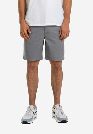 Hombre con pantalones cortos grises hasta la rodilla, camiseta blanca y zapatillas Nike Air Max blancas, de pie frente a un fondo blanco liso.