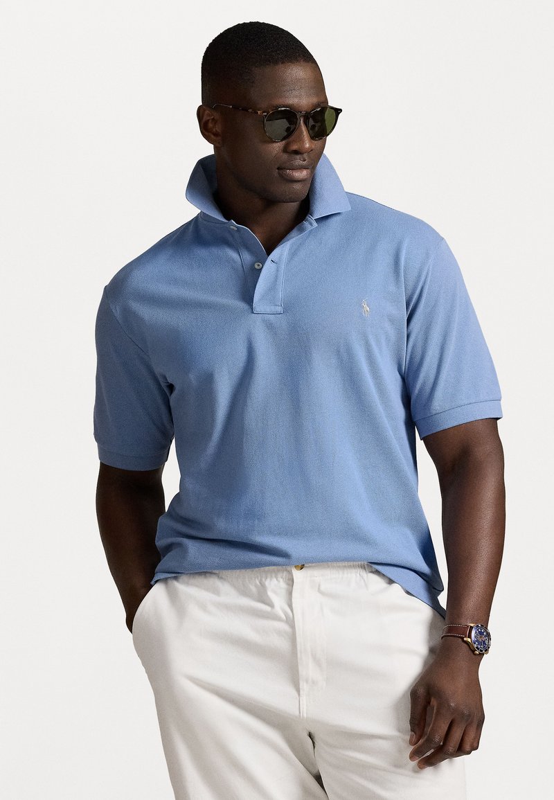 Camisa polo azul clara com colarinho estruturado, mangas curtas e um logotipo bordado. Combinada com calções brancos e um relógio castanho.