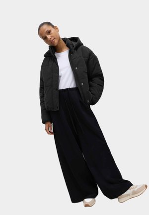 Chaqueta de plumas negra con capucha, textura acolchada y botones a presión, combinada con pantalones negros de corte holgado y zapatillas beige. Camiseta blanca por debajo.