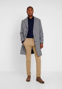 Płaszcz w wzór pepitki w czerni i bieli, zestawiony z granatową koszulą i beżowymi spodniami, uzupełniony brązowymi botkami za kostkę.