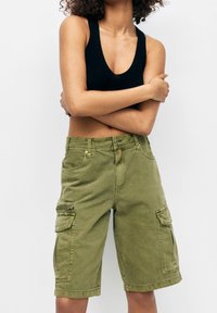 Person iført olivengrønne cargo shorts og en sort ærmeløs cropped top, med armene krydset over torsoen foran en ensfarvet baggrund.