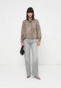 Blusa leopardata con colletto annodato, abbinata a jeans grigi chiari a zampa d'elefante. Borsa nera, tacchi nudi, sfondo minimalista.