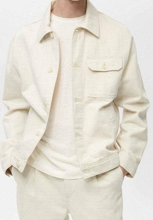 Licht beige denim jack met een knoopsluiting aan de voorkant, een enkele borstzak en een klassieke kraag; gedragen over een eenvoudig licht topje. Soepele textuur.