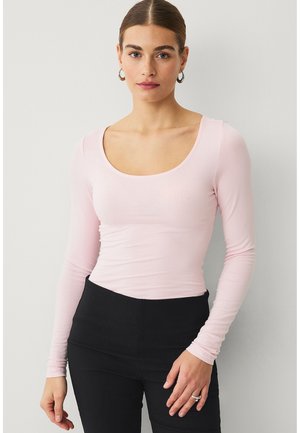 Kvinde med håret trukket tilbage, iført lyserød top med lange ærmer og rund hals samt sorte bukser, stående mod en lys grå væg.