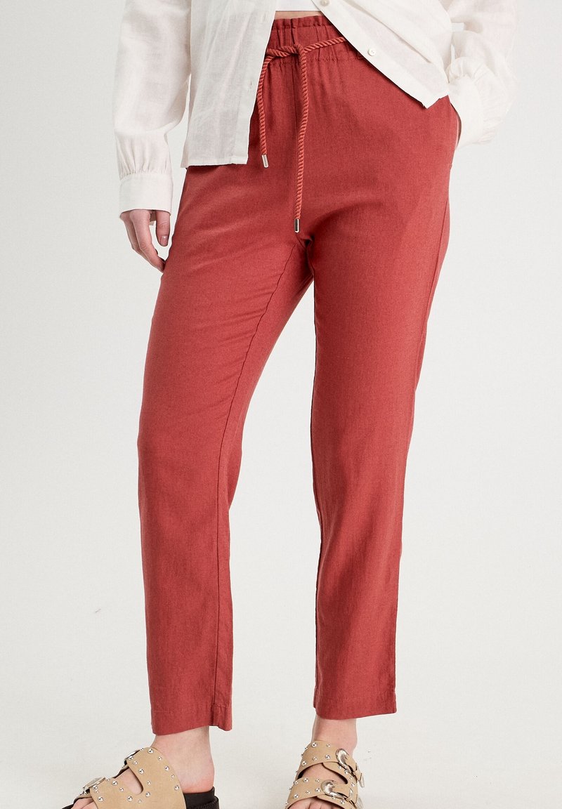 Personne portant un pantalon à cordon rouille, des sandales beiges cloutées, et une chemise à manches longues beige clair aux manches retroussées.