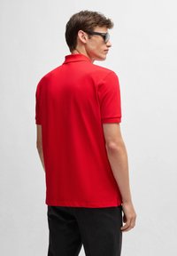 Röd polo skjorta med en klassisk krage och korta ärmar. Tillverkad av ett mjukt tyg. Svarta byxor. Modell sedd bakifrån, bärande solglasögon.