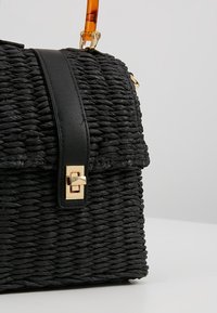 Sac à main noir tissé avec une bandoulière en cuir et une fermeture à torsion dorée, avec une poignée courte couleur ambre sur un fond gris uni.