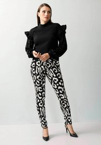 Schwarzes Top mit gerafften, schwarzen Paillettenärmeln, kombiniert mit schwarzen und weißen Leopardenmuster-Hosen. Model trägt schwarze spitze Absatzschuhe.