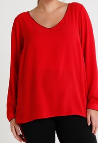 Kobieta w luźnej, czerwonej bluzce z długim rękawem i dekoltem w serek oraz czarnych spodniach, przedstawiona od szyi do bioder.