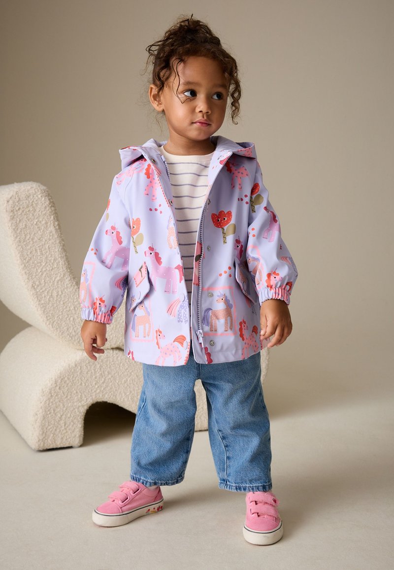 Jeune enfant portant une veste violet clair avec des licornes et des fleurs, un t-shirt rayé, un jean bleu et des baskets roses, debout à l'intérieur.