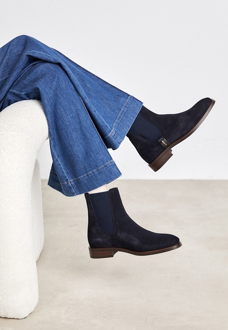 Personne portant des bottines Chelsea en daim bleu foncé et un jean large bleu, assise sur une chaise blanche texturée sur un fond uni.