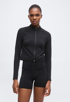 Femme portant une veste noire zippée à manches longues et un short de sport noir, debout devant un fond clair uni.