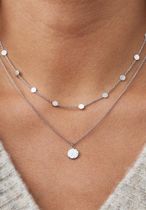 Deux colliers en argent, l'un avec des disques ronds espacés et l'autre avec un pendentif circulaire texturé, reposant sur la peau nue.