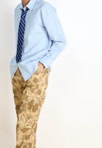 Jasnoniebieska koszula na guziki z ostrym kołnierzem, zestawiona z brązowymi spodnie bojówkami w kamuflaż, wyposażonymi w wiele kieszeni i teksturowaną tkaninę.