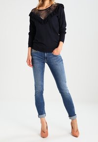 Mujer vestida con una blusa negra de manga larga con encaje y volantes, unos jeans ajustados azul con puños, y zapatos de tacón melocotón, de pie contra un fondo blanco.